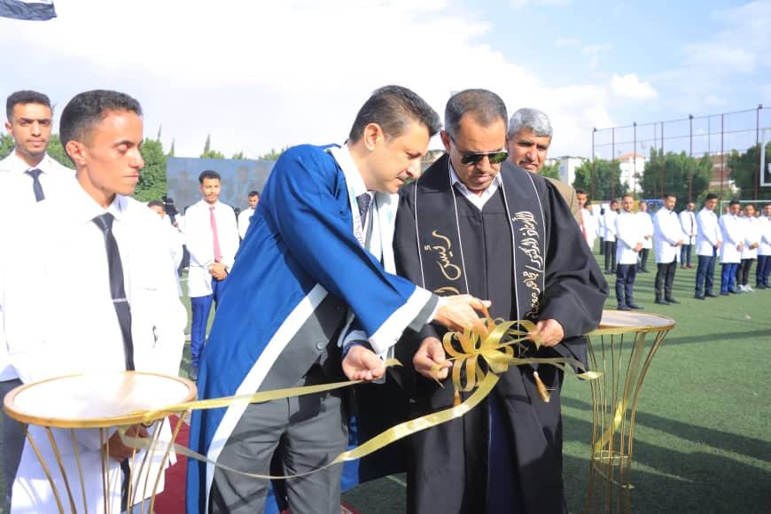 1003982547 - جامعة 21 سبتمبر Two men in ceremonial robes cut a gold ribbon at a ribbon-cutting on a sports field, with students watching in white shirts and ties in the background.