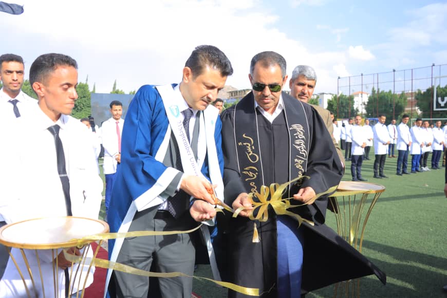 1003982550 - جامعة 21 سبتمبر Two men in academic robes cut a gold ribbon at an outdoor graduation ceremony, with students in white coats lined up in the background.