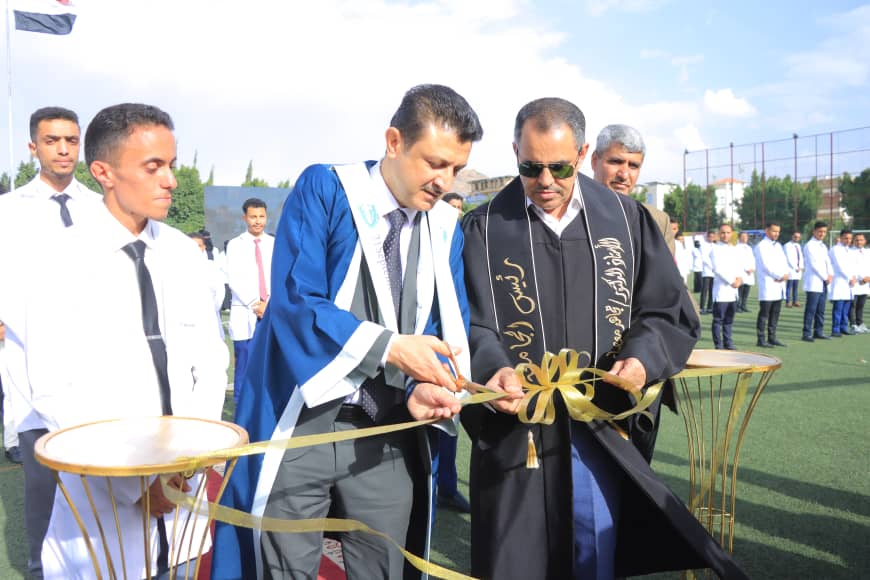 1003982553 - جامعة 21 سبتمبر Two men in ceremonial robes cut a gold ribbon at an outdoor ribbon-cutting ceremony with students lined up behind them on a sports field.