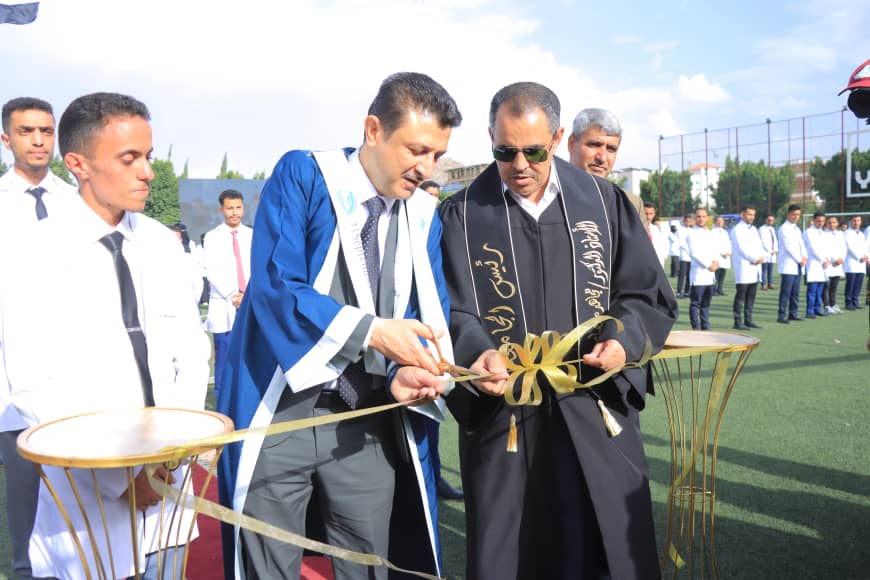 1003982556 - جامعة 21 سبتمبر Two men in ceremonial robes cut a gold ribbon at an outdoor event on a sports field, with teams of students in white shirts in the background.