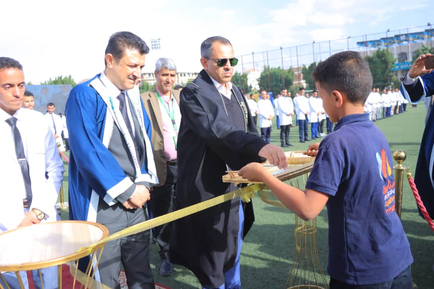 1003982559 - جامعة 21 سبتمبر Official in dark robe hands a small wrapped item to a boy at a ceremonial award on a sports field, with spectators and line of participants in the background.