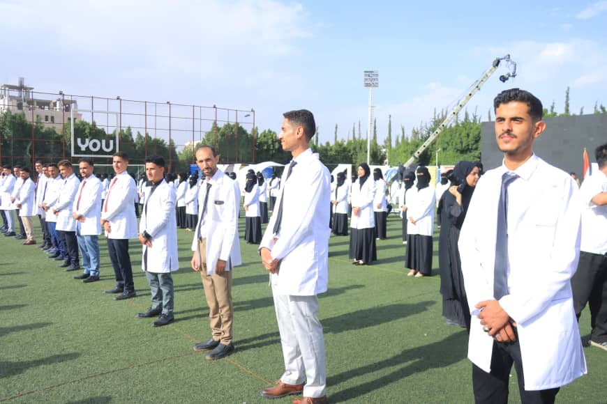 1003982562 - جامعة 21 سبتمبر Line of people in white lab coats standing on a sports field during a ceremony, with others in the background and a clear blue sky.