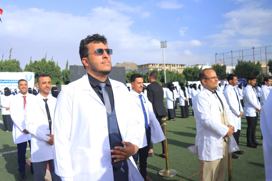 1003982565 - جامعة 21 سبتمبر Young men in white lab coats stand in an outdoor field during a formal ceremony; foreground man wears sunglasses and a dark suit.