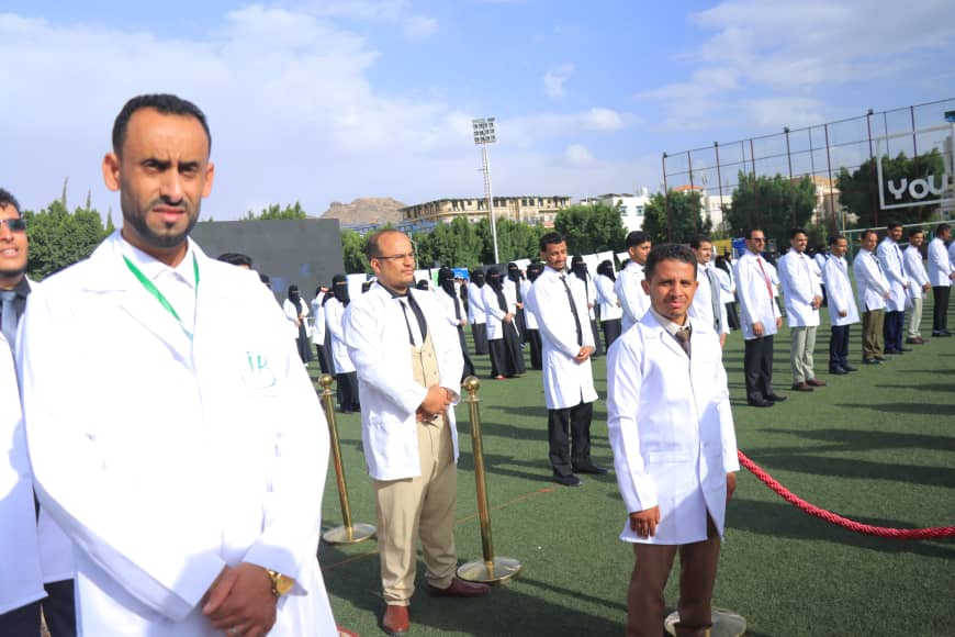 1003982568 - جامعة 21 سبتمبر Group of doctors in white coats lined up on a sports field during an outdoor ceremony; two men in foreground face the camera.