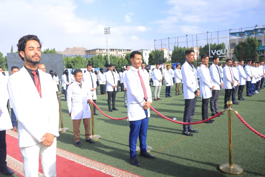 1003982571 - جامعة 21 سبتمبر Line of people in white lab coats standing behind a red rope on a grassy ceremonial field, likely a student or medical event.