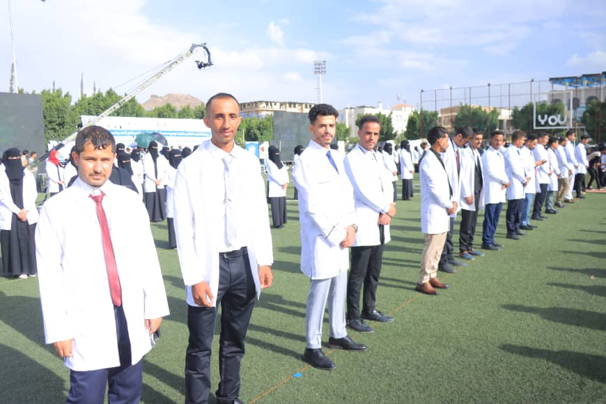 1003982574 - جامعة 21 سبتمبر Line of people in white coats standing in a row on a grassy field during an outdoor event; some wear ties, others in traditional dress in the background.