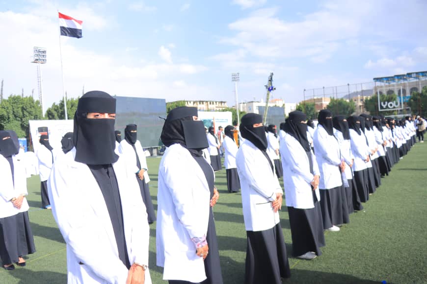 1003982577 - جامعة 21 سبتمبر Line of women in white lab coats and black niqabs standing in formation on a sports field, Egyptian flag in background.