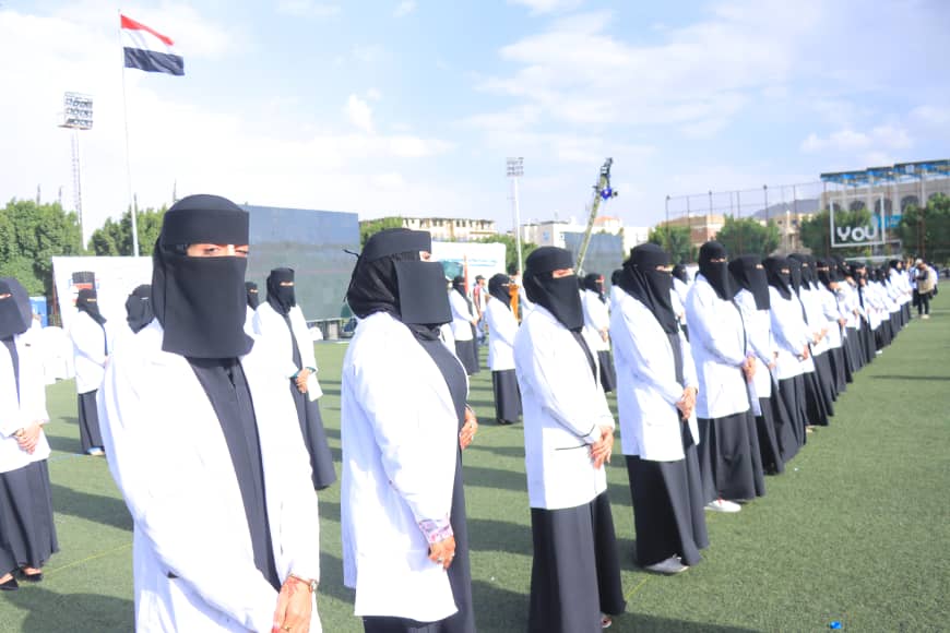 1003982580 - جامعة 21 سبتمبر Line of women in white coats and black niqabs standing at attention on a grassy sports field during an outdoor event, flag in background.