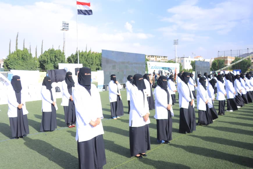 1003982583 - جامعة 21 سبتمبر Line of women in white lab coats and black niqabs standing in rows on a sports field during an outdoor ceremony, Egyptian flag in the background