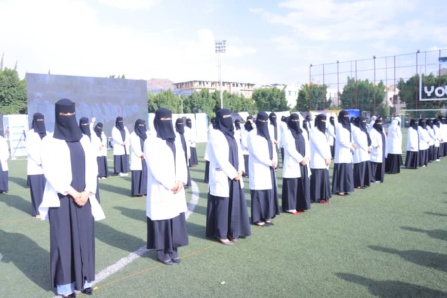 1003982586 - جامعة 21 سبتمبر Rows of women in black niqabs and white lab coats standing on a sports field for a formal event or ceremony, with a clear blue sky in the background.