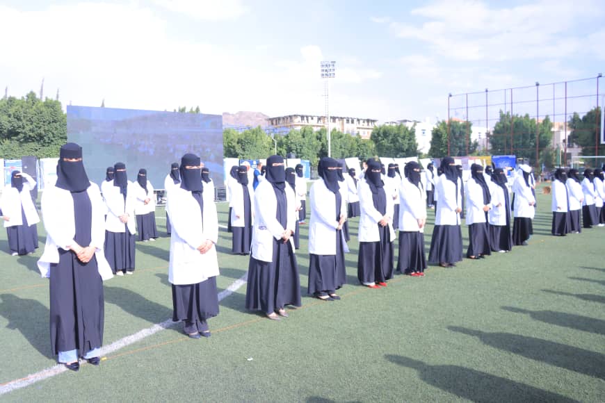 1003982589 - جامعة 21 سبتمبر Group of women wearing black veils and white lab coats standing in neat rows on a grassy sports field outdoors, socially distanced.