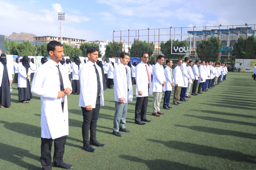 1003982592 - جامعة 21 سبتمبر Group of students in white lab coats standing in neat rows on a grassy field during an outdoor ceremony or drill, with buildings and bleachers in the background on a sunny day.