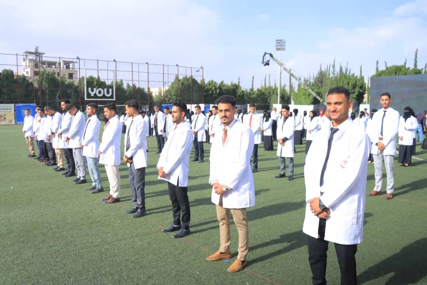 1003982595 - جامعة 21 سبتمبر Group of medical students in white lab coats lined up on a sports field during an outdoor ceremony, with a 'YOU' banner in the background.
