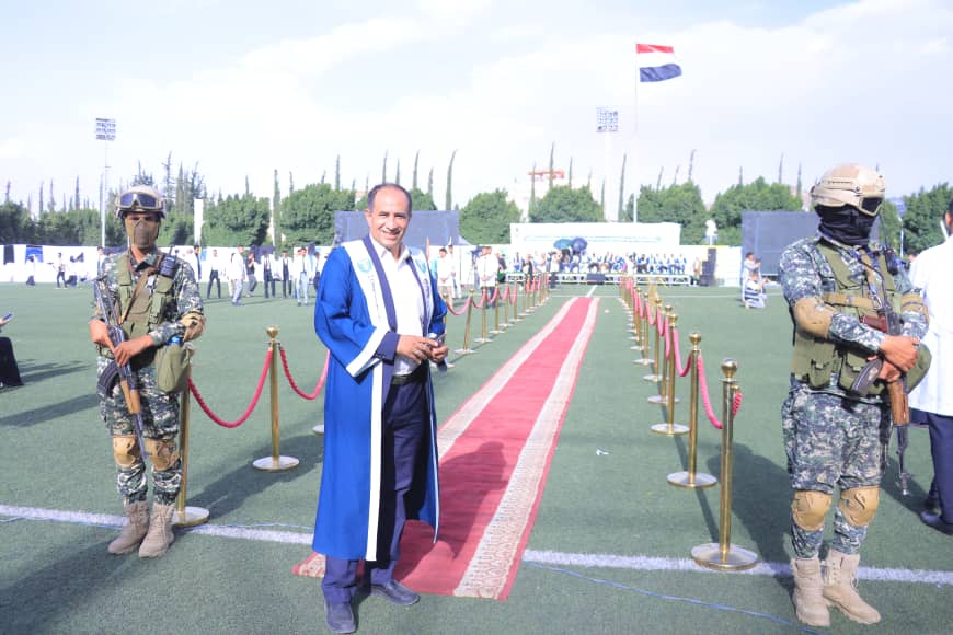 1003982598 - جامعة 21 سبتمبر Man in a blue ceremonial robe walking on a red carpet flanked by armed guards at an outdoor event on a sports field.