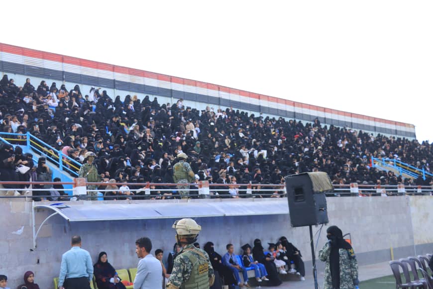 1003982601 - جامعة 21 سبتمبر Large crowd seated in stadium bleachers during an outdoor event, with security personnel at the front.