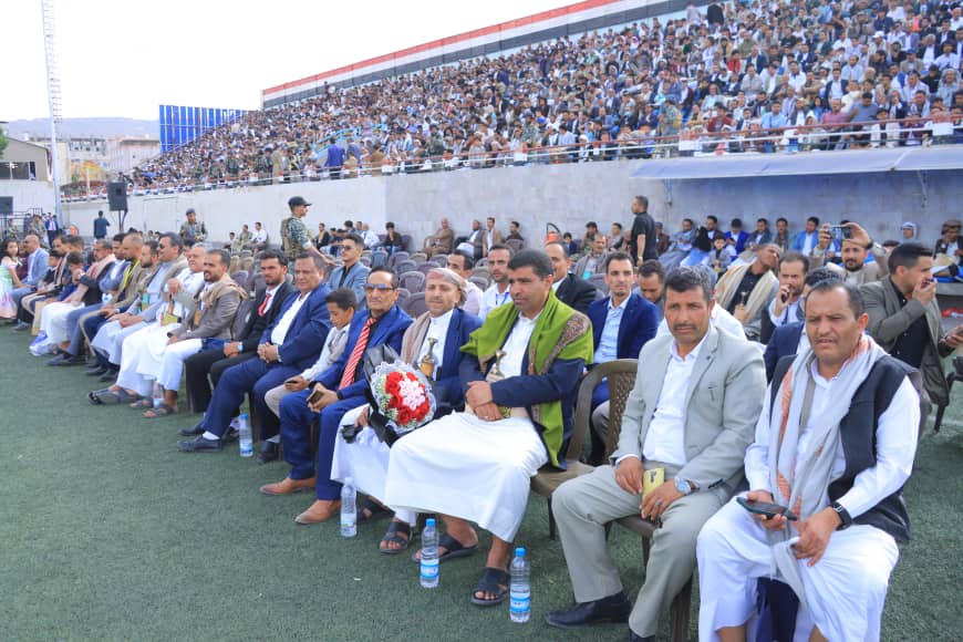 1003982607 - جامعة 21 سبتمبر Rows of men in formal and traditional dress seated on a sports field during a ceremony, with a packed stadium stands behind them under bright light.