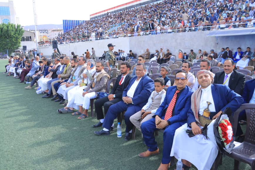 1003982610 - جامعة 21 سبتمبر Formal audience seated in stadium seats for a ceremony, with a large crowd in the background.