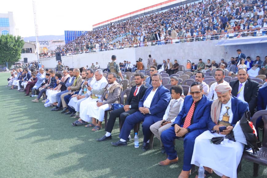 1003982613 - جامعة 21 سبتمبر Rows of formally dressed attendees seated in chairs on a grassy field at an outdoor stadium, with a large audience in the stands behind them.