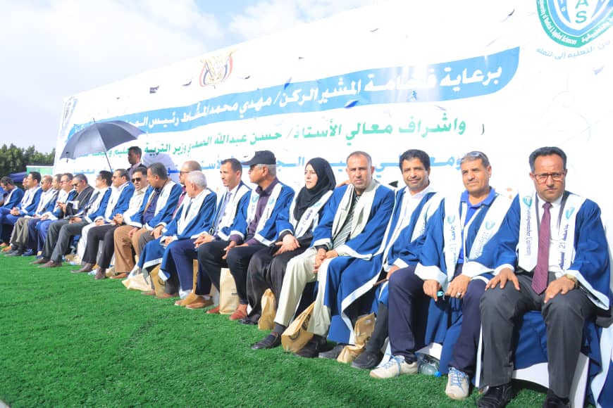 1003982616 - جامعة 21 سبتمبر Row of people in blue academic robes seated on a lawn during an outdoor ceremony with a large Arabic banner behind them.