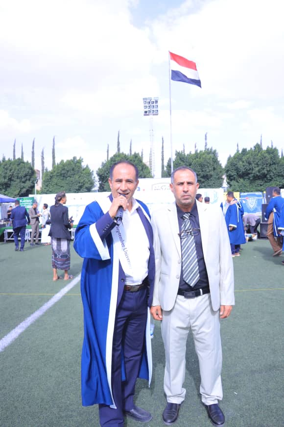 1003982619 - جامعة 21 سبتمبر Two men at a sports field: one in a blue gown holding a microphone, the other in a light gray suit, with a Dutch flag in the background.