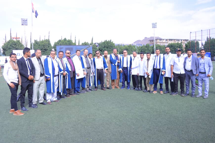1003982622 - جامعة 21 سبتمبر Group of people standing in a line on a sports field, some wearing blue academic gowns and scarves, posing for a ceremony photo.