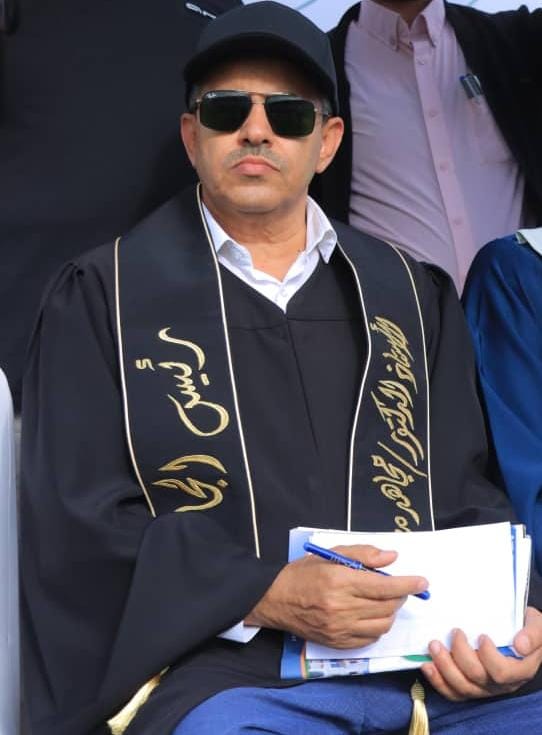 1003983803 - جامعة 21 سبتمبر Man in a black graduation gown and cap with a gold-embroidered stole, wearing sunglasses and holding papers with a blue pen.