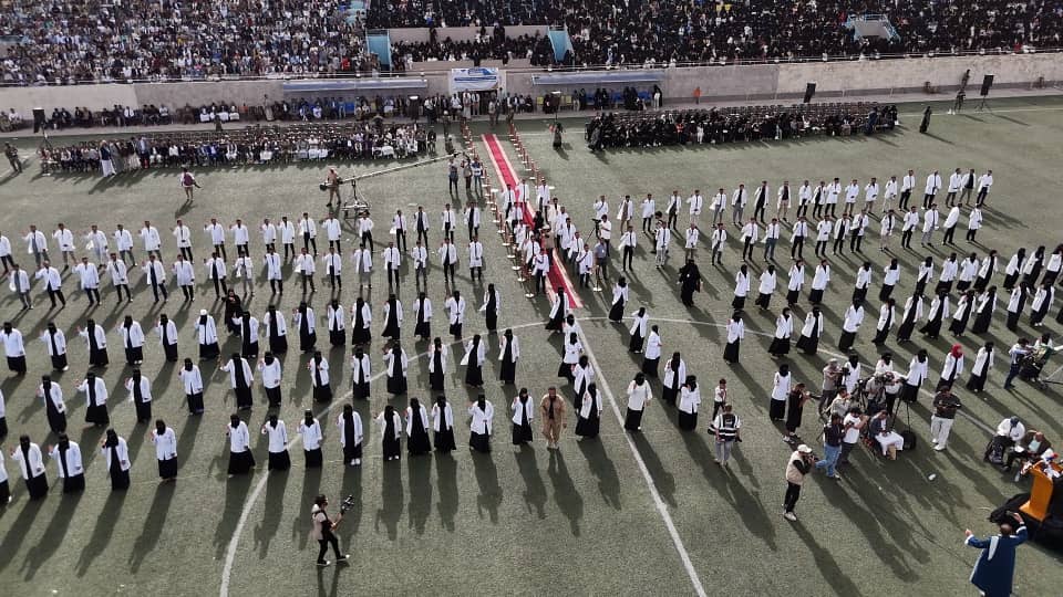 1003984138 - جامعة 21 سبتمبر Aerial view of a large ceremony on a sports field with hundreds of students in white shirts and dark skirts arranged in diagonal lines, facing inward toward a red carpet centerpiece, with spectators in stands around the field somewhat blurred in the background.
