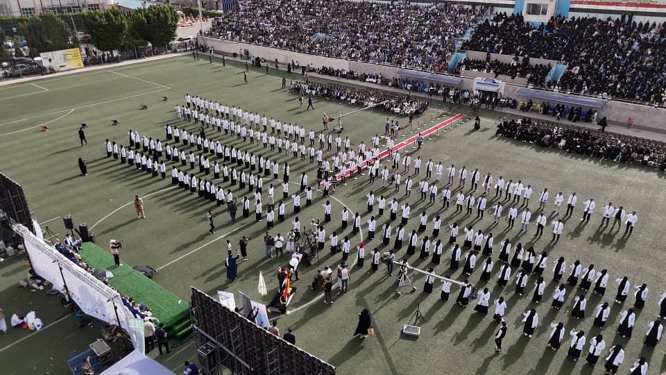 1003984141 - جامعة 21 سبتمبر Aerial view of a stadium field with hundreds of people in white uniforms forming organized rows and shapes, while large crowds watch from the stands.