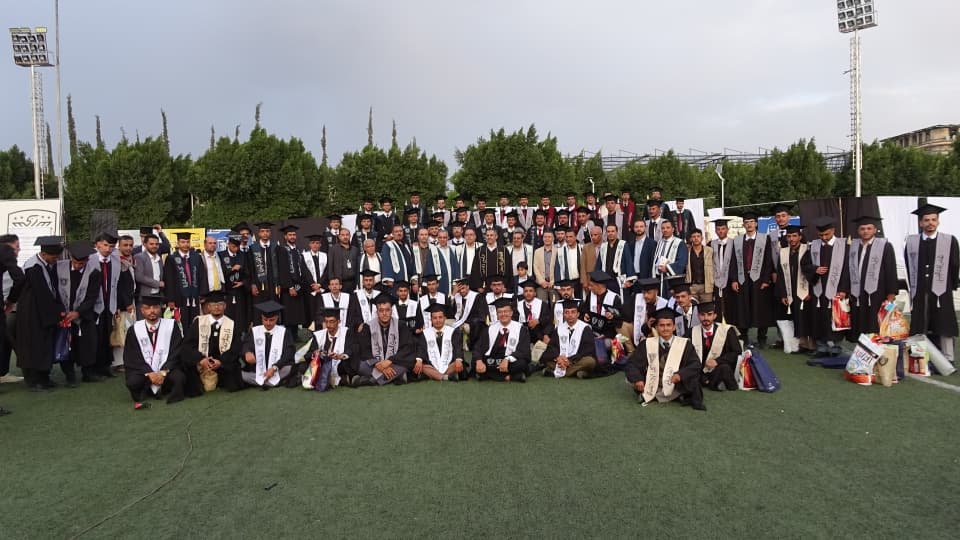 1003984188 - جامعة 21 سبتمبر Large group of graduates in caps and gowns posing on a sports field, with stoles and gift bags nearby, trees in the background.
