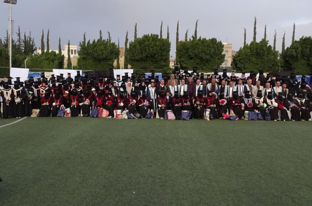 1003984191 - جامعة 21 سبتمبر Large group of graduates and faculty in gowns posing on a soccer field during a graduation ceremony, with trees in the background.