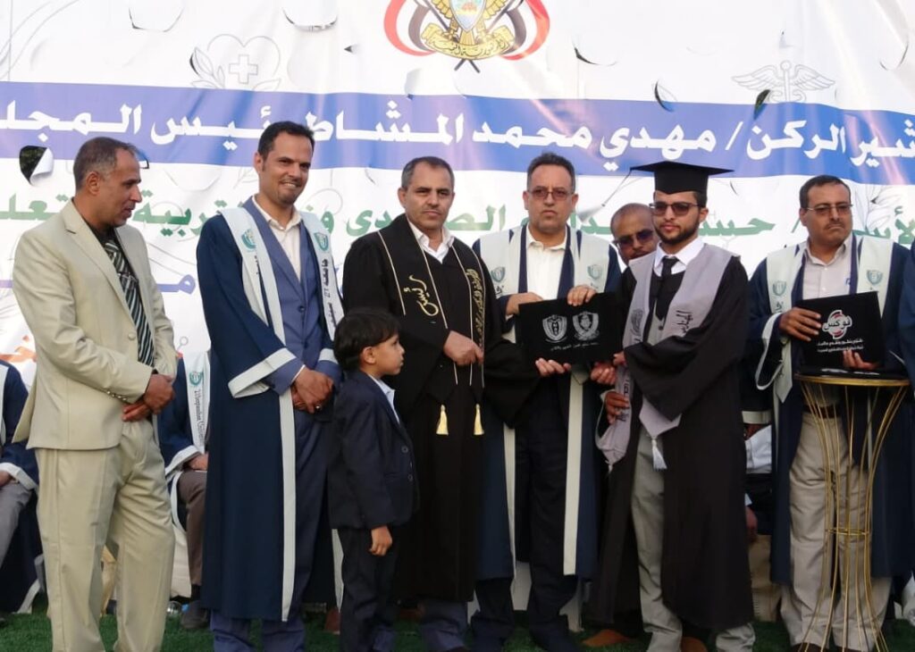 1003984194 - جامعة 21 سبتمبر Graduates in blue and white robes on stage during a ceremony, with a graduate receiving a diploma as a small boy stands nearby.