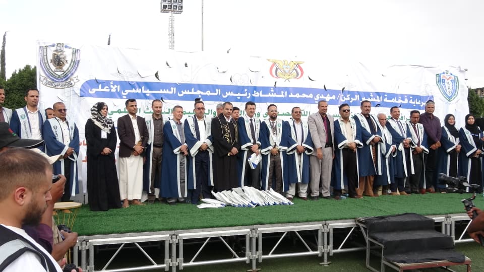 1003984197 - جامعة 21 سبتمبر Group of graduates in blue robes posing on a stage during a ceremony, with a large Arabic banner behind them and diplomas on the ground in front of the line of people.