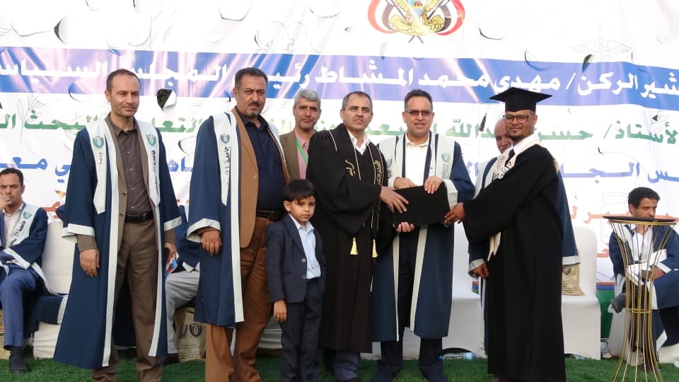 1003984209 - جامعة 21 سبتمبر Group of graduates and officials posing on stage at a graduation ceremony; a man in a black gown holds a diploma while others smile, with a banner in Arabic behind them and a young boy standing in front.