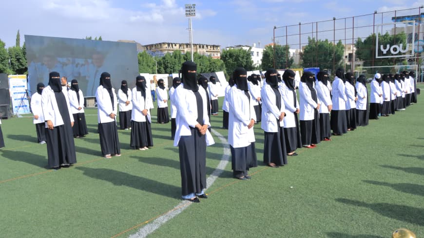 1003984220 - جامعة 21 سبتمبر Group of women in black abayas and white lab coats standing in neat rows on a green sports field during an outdoor ceremony.