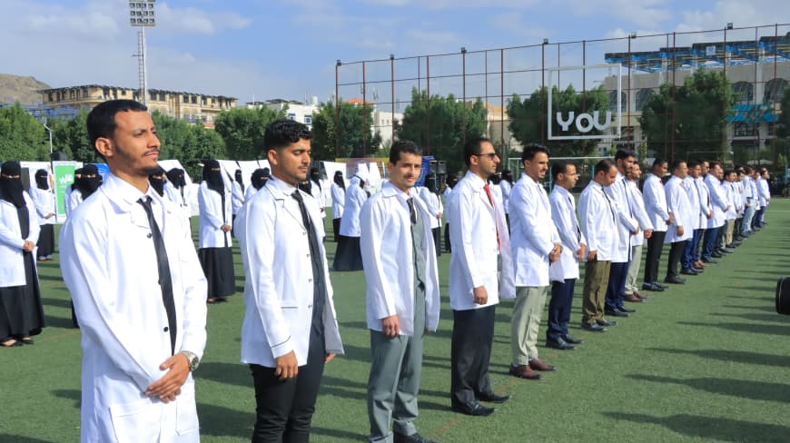 1003984223 - جامعة 21 سبتمبر Group of people in white lab coats standing in a straight line on a sports field during an outdoor event.