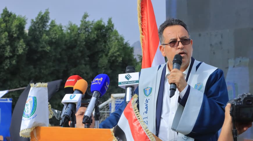 1003984229 - جامعة 21 سبتمبر Public speaker at an outdoor press conference, standing behind a podium with multiple microphones and flags in the background.