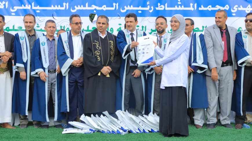1003984232 - جامعة 21 سبتمبر Group of graduates in blue and white robes on a stage outdoors; a woman hands a certificate to a man amid a banner with Arabic text.