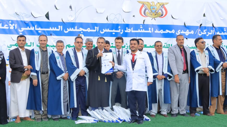 1003984235 - جامعة 21 سبتمبر Group of men in robes on a stage receiving certificates at an outdoor ceremony with a blue banner in Arabic in the background.