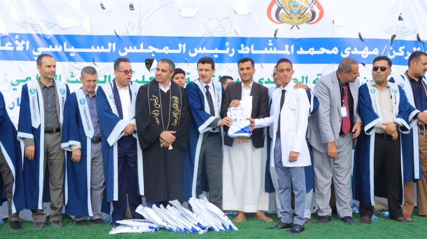 1003984238 - جامعة 21 سبتمبر Group of men in blue and white academic gowns stand on a stage as an award is presented, with an Arabic banner in the background.