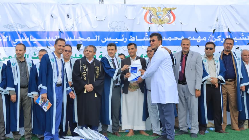 1003984241 - جامعة 21 سبتمبر Group of men in blue and white ceremonial gowns pose together on a stage during an award or graduation ceremony, with a blue banner and national emblem behind them.