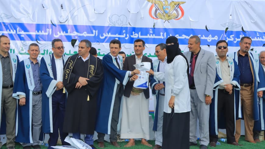 1003984244 - جامعة 21 سبتمبر Group of people in blue ceremonial robes stand on a stage while a woman hands a document to a man during a formal ceremony, banner with Arabic text behind them