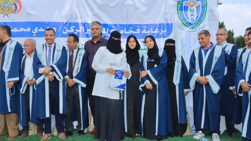 1003984247 - جامعة 21 سبتمبر Group of people in blue graduation gowns standing outdoors for a ceremony, with a banner and logos in the background.