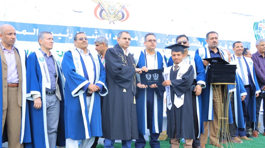 1003984250 - جامعة 21 سبتمبر Graduate in cap and gown receives diploma from faculty member during a ceremony; group of men in blue robes on stage with a blue banner in Arabic in the background.