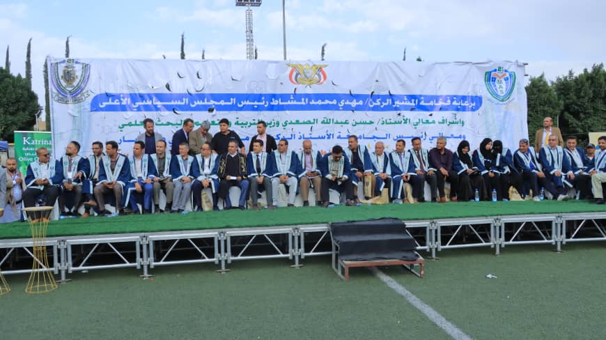 1003984313 - جامعة 21 سبتمبر Group of men and women seated on a stage outdoors during a ceremony, wearing blue sashes, with a large Arabic banner and crests in the background.