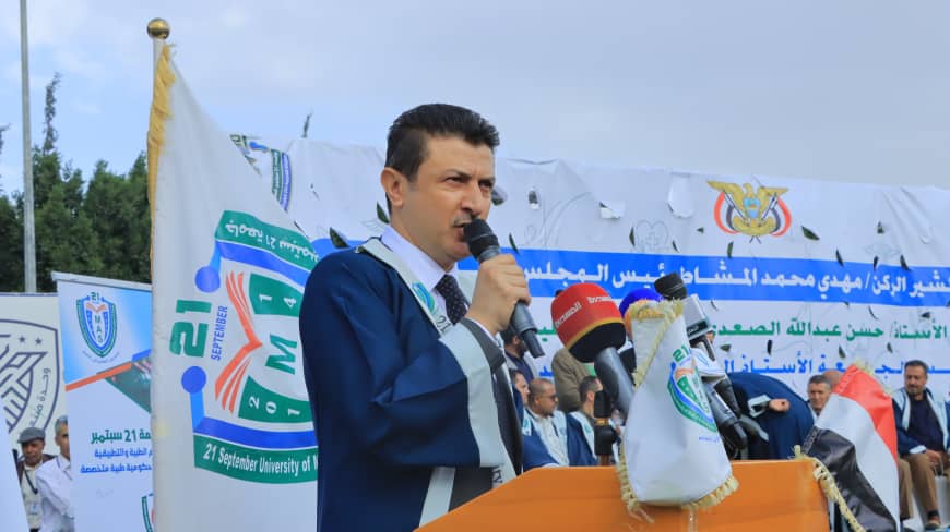 1003984316 - جامعة 21 سبتمبر Man in a blue gown speaks into a microphone at a podium during a university event, with banners and a crowd in the background.