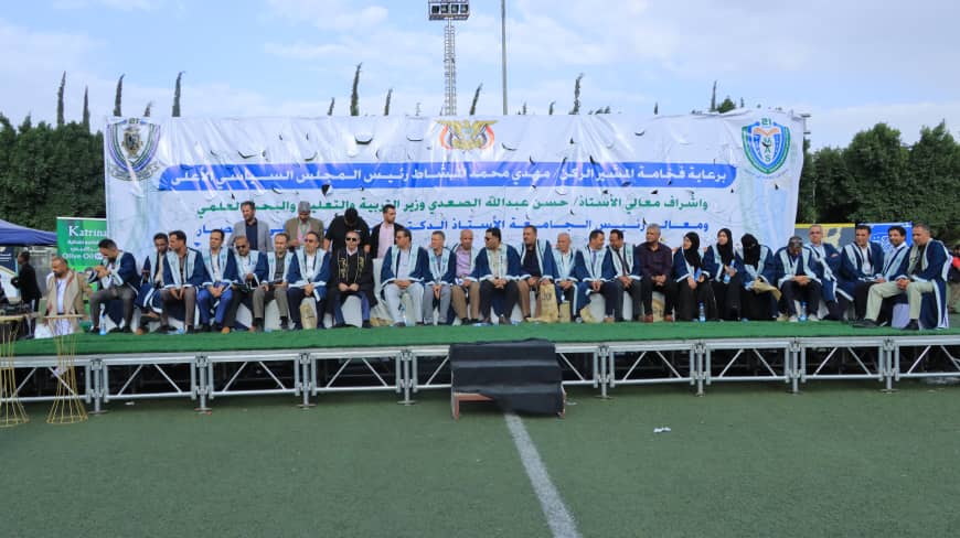 1003984319 - جامعة 21 سبتمبر Group of graduates or officials seated on a stage in front of a large blue banner at an outdoor ceremony on a sports field.
