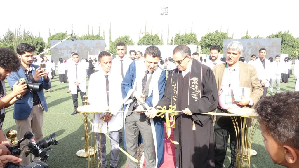 1003984322 - جامعة 21 سبتمبر Group of graduates and faculty cutting a ribbon at an outdoor ceremony on a green field, with photographers capturing the moment.