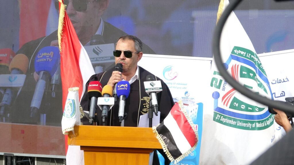 1003984323 - جامعة 21 سبتمبر Man in sunglasses speaking into a microphone at a podium during a press conference, with multiple flags and microphones in front
