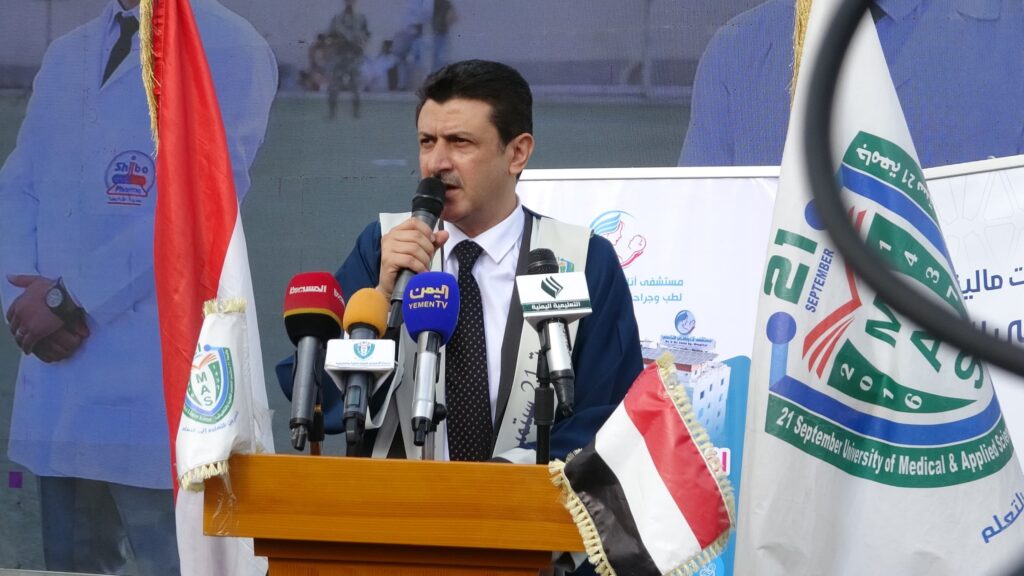 1003984324 - جامعة 21 سبتمبر Man in a suit speaks into a handheld microphone at a podium, with multiple microphones, flags, and banners behind him at a public event.
