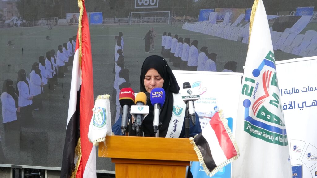 1003984325 - جامعة 21 سبتمبر Woman in a black hijab speaks at a podium with multiple microphones, flanked by flags during an outdoor event. Behind her is a large screen showing people on a field.